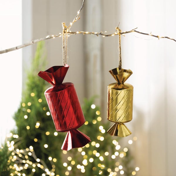 Zwei als Bonbons gestaltete Weihnachtsanhänger hängen an einem Ast vor einem beleuchteten Tannenbaum.