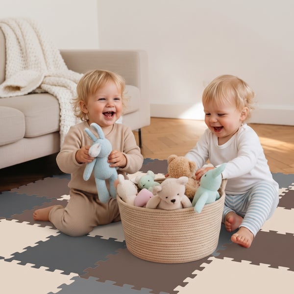 Zwei Kleinkinder spielen auf einer puzzleartigen Bodenmatte aus Schaumstoff mit Kuscheltieren in einem Korb.