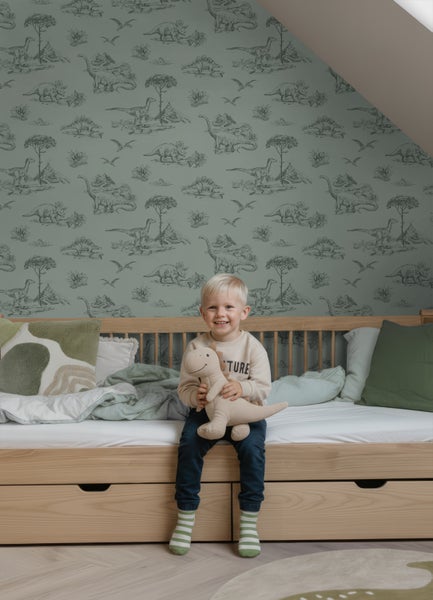 Vliestapete mit Dinosaurier-Motiven in einem hellen Kinderzimmer. Ein lächelndes Kind sitzt auf einem Holzbett vor der gemusterten Wand.