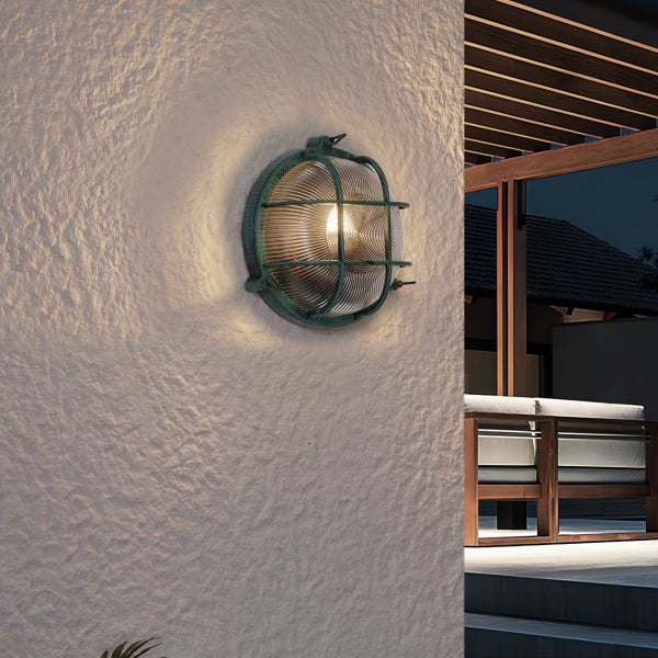 Runde maritime Außenwandleuchte mit Schutzgitter an einer weißen Putzfassade neben einer beleuchteten Terrasse.