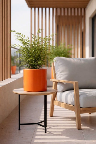 Zylindrischer Pflanztopf mit Pflanze auf einem runden Holztisch neben einem Gartenstuhl auf einem hellen, modernen Balkon.