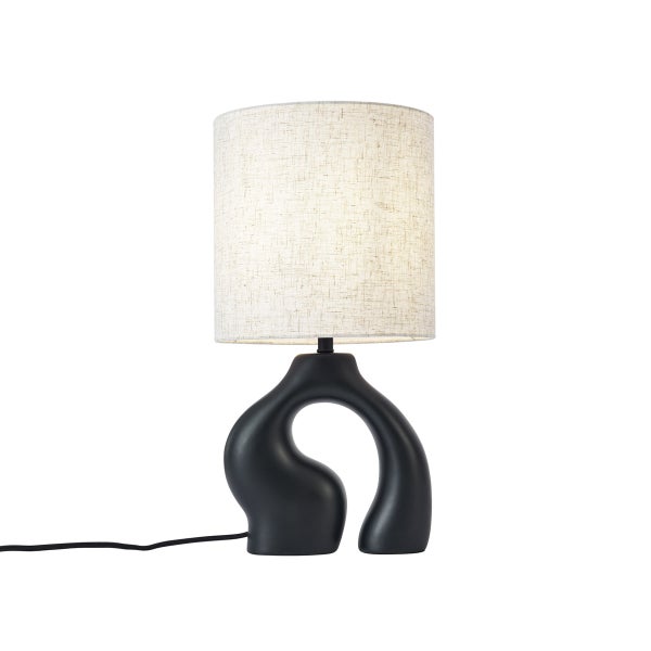 Tischleuchte mit abstraktem schwarzem Fuß und zylindrischem beigem Textilschirm.
