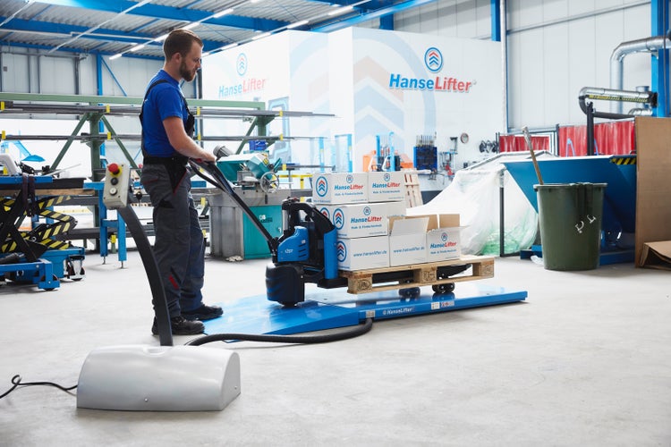 Mitarbeiter bedient elektrischen Hubwagen mit Wiegefunktion auf einer Bodenwaage in einem Lager mit HanseLifter Logo und Versandkartons auf Palette.
