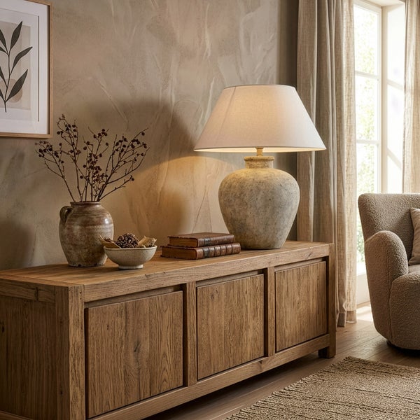 Rustikales Sideboard aus Holz dekoriert mit einer Tischlampe aus Keramik, einer Vase mit Zweigen und Büchern in einem hellen Wohnzimmer.