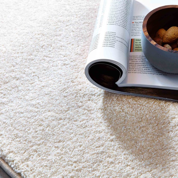 Hochflor-Teppich in Creme mit weicher, zotteliger Textur, darauf liegen ein Magazin und eine Holzschale mit Nüssen.