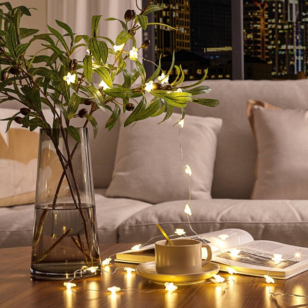 LED-Lichterkette mit blumenförmigen Leuchten, dekoriert an einem Olivenzweig in einer Glasvase und auf einem Tisch neben einer Tasse und einem Buch.