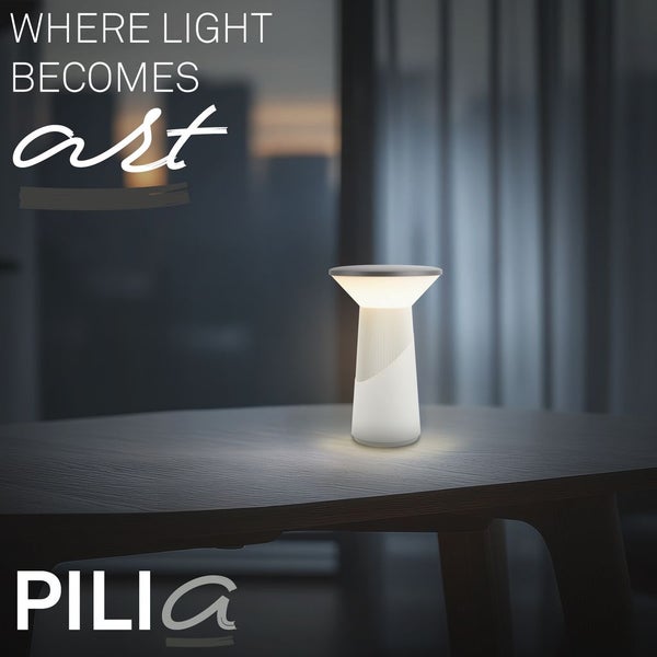 Moderne Pilia Tischleuchte auf einem Holztisch in einer atmosphärisch beleuchteten Szene. Pilia Logo. Text Where light becomes art.