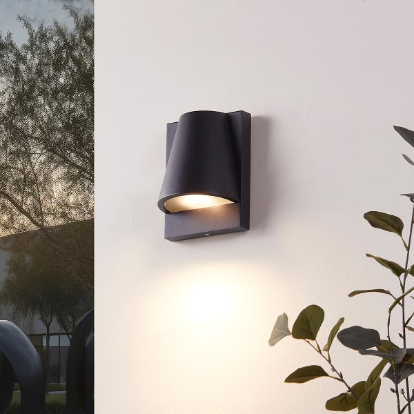 Moderne schwarze Außenwandleuchte aus Metall an einer hellen Wand, kegelförmiges Gehäuse mit warmem Lichtstrahl nach unten, im Hintergrund Pflanzen.