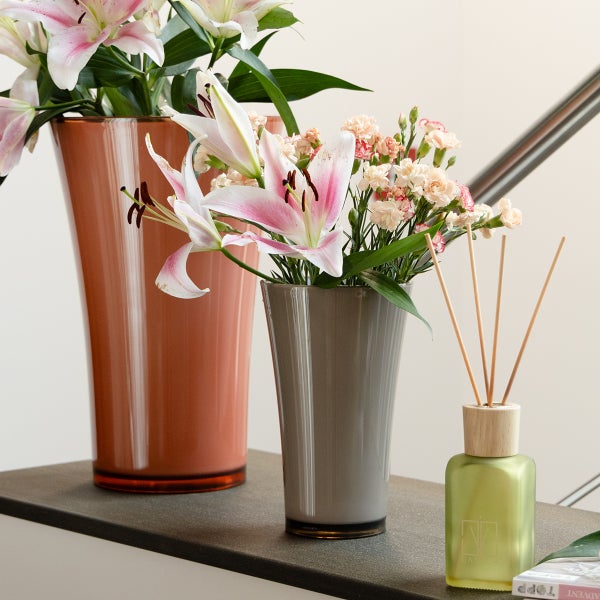Zwei Glasvasen in Grau und Orange mit Lilien und Nelken neben einem Raumduft Diffusor auf einer Ablage.