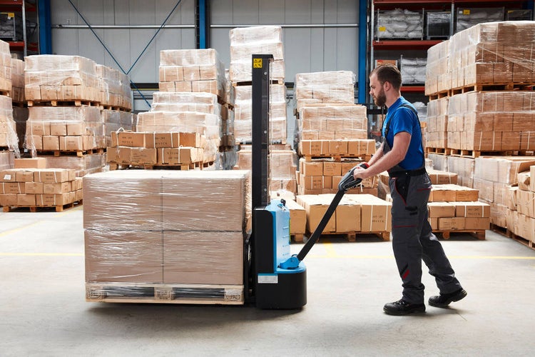 Mann bedient einen Elektro-Hochhubwagen in einem Lagerhaus, um eine Palette mit Kartons zu transportieren.