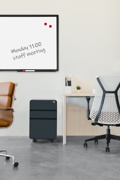 Modernes Büro mit Whiteboard, schwarzem Rollcontainer aus Metall, hellem Schreibtisch und ergonomischen Bürostühlen in einem hellen Raum.