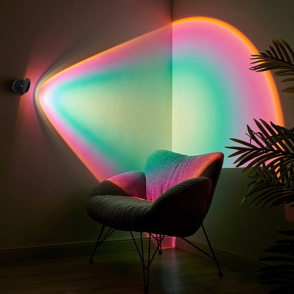An der Wand montierte LED-Projektionslampe strahlt mehrfarbiges Licht in eine Zimmerecke über einem grauen Sessel und einer Zimmerpflanze.