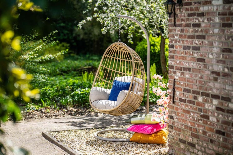 Hängesessel in Eiform aus Geflecht mit Gestell und Polstern in einem hellen Garten neben einer Backsteinwand.
