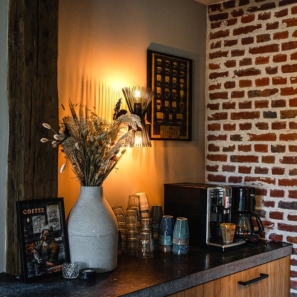 Küchenecke mit Kaffeemaschine, Gläsern, Trockenblumen in Vase, Wandleuchte und gerahmten Bildern