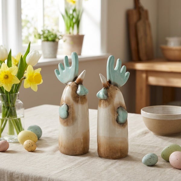 Zwei dekorative Keramikfiguren in Form von Hähnen auf einem Tisch mit Osterdekoration und Blumen in einer Glasvase.