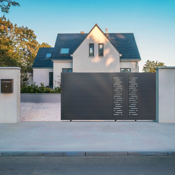 Modernes anthrazitfarbenes Schiebetor aus Aluminium vor einem Wohnhaus mit weißer Fassade und Kiesauffahrt.