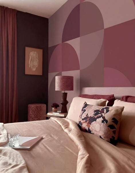 Schlafzimmer mit geometrischer Wandgestaltung in Beere-Tönen, Bett mit heller Bettwäsche und floralem Kissen sowie Nachttischlampe.