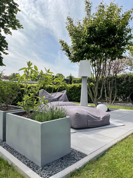 Moderne Gartenlounge mit grauen rechteckigen Pflanzkübeln, einem lila Sitzsack und einer hohen Außenstehleuchte auf einer hellen Steinterrasse.