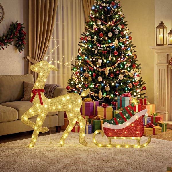 Beleuchtete Weihnachtsdekoration mit Rentier und Schlitten vor einem geschmückten Weihnachtsbaum und Geschenken im Wohnzimmer.