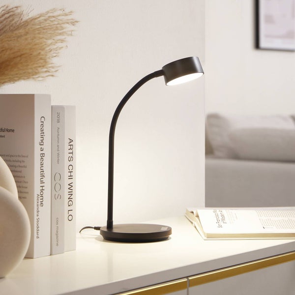 Moderne schwarze LED-Schreibtischlampe mit gebogenem Hals und rundem Standfuß auf einer weißen Oberfläche neben Büchern.