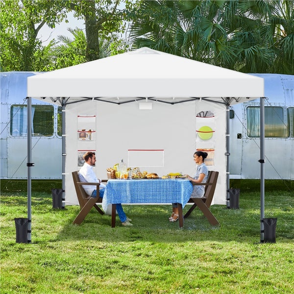 Szene mit einem weißen Faltzelt, unter dem ein Paar an einem mit blau karierter Tischdecke gedeckten Tisch sitzt