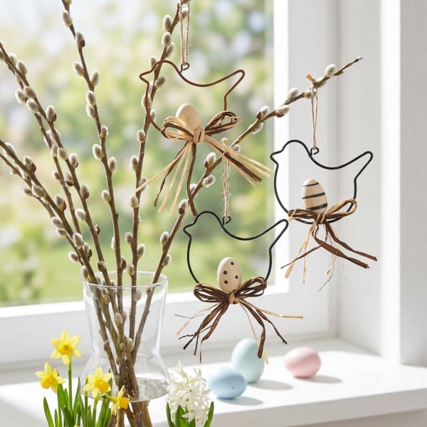 Osterdekoration zum Hängen in Vogelform mit kleinen Ostereiern und Bastschleife an Weidenzweigen vor einem Fenster.