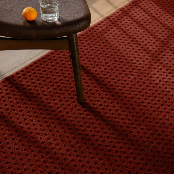 Nahaufnahme eines strukturierten Teppichs mit Punktmuster auf einem Holzfußboden, neben einem Hocker mit einem Wasserglas und einer Frucht.