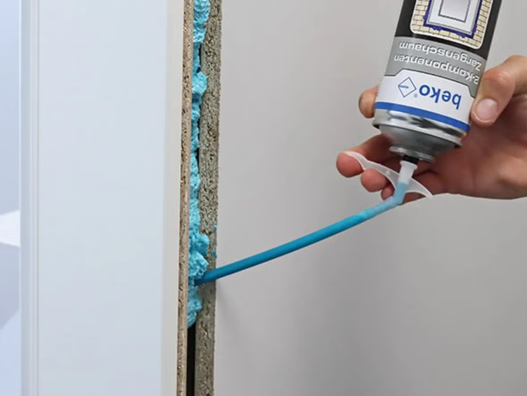 Eine Hand trägt mit einer Beko Zwei Komponenten Zargenschaum Sprühdose Montageschaum in einen Türzargenspalt auf.