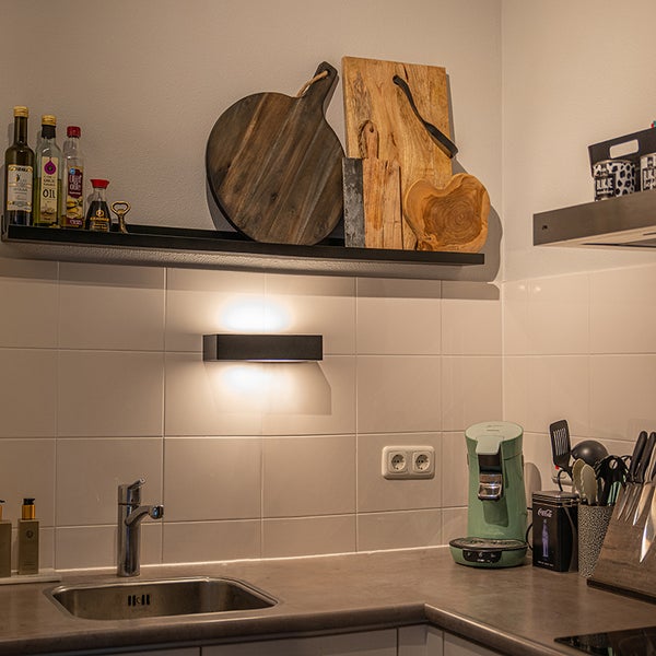 Küchenszene mit Spüle, Armatur, Wandleuchte, Regalen mit Dekoration und Küchenutensilien.