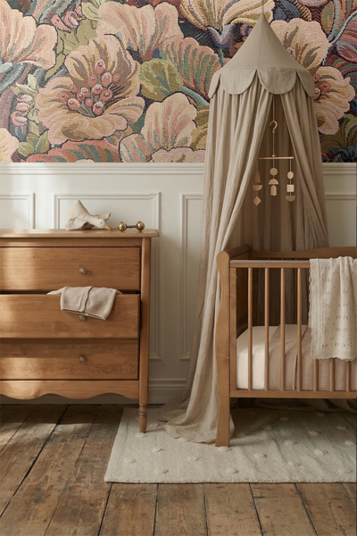 Helles Kinderzimmer mit Holzkommode und passendem Gitterbett mit Betthimmel vor einer floralen Tapete und weisser Wandtäfelung.