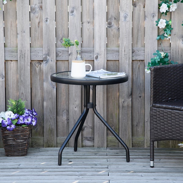 Runder Bistrotisch mit Glasplatte und schwarzem Metallgestell auf einer Holzterrasse mit einem Korbstuhl und Blumen in einer organisierten Umgebung.