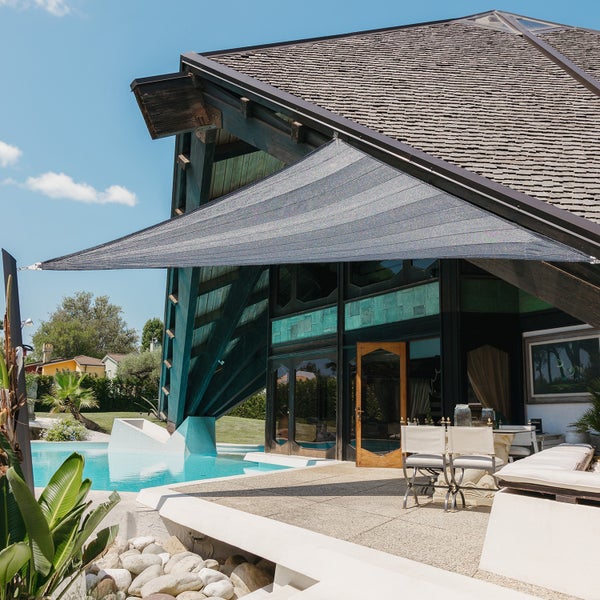 Dreieckiges Sonnensegel über einer modernen Terrasse mit Pool und Gartenmöbeln vor einem dunklen Holzhaus.