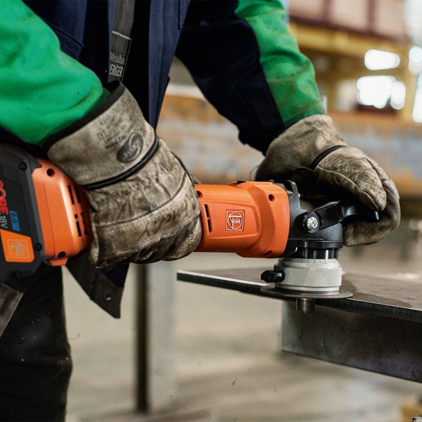 Person in Arbeitshandschuhen bedient eine orangefarbene Fein akkubetriebene Kantenfräse an einer Metallplatte in einer Werkstatt.