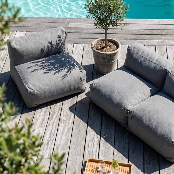 Graue Outdoor-Lounge-Module auf einer Holzterrasse am Pool mit einem Olivenbaum im Topf in einer hellen Atmosphäre.