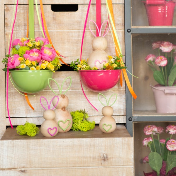 Dekorative Szene mit hängenden Pflanzgefäßen, Blumenarrangements, Schleifenbändern und hölzernen Hasenfiguren auf einer Holzoberfläche
