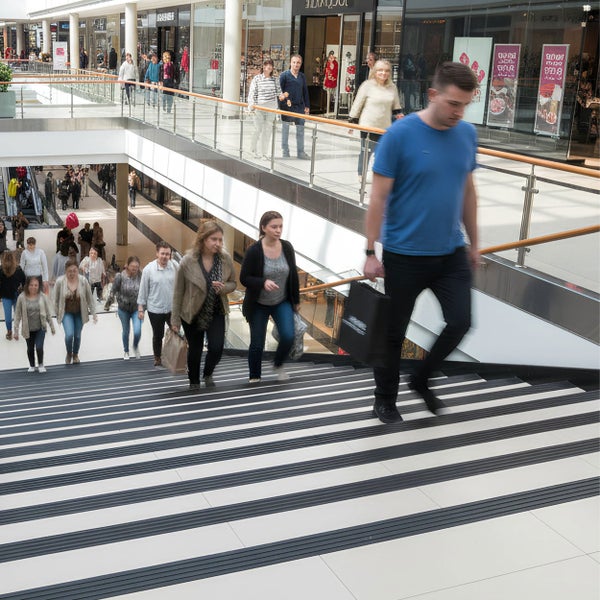 Moderne Treppe in einem Einkaufszentrum mit schwarzen Antirutschstreifen auf hellen Stufen und Personen, die sie benutzen.
