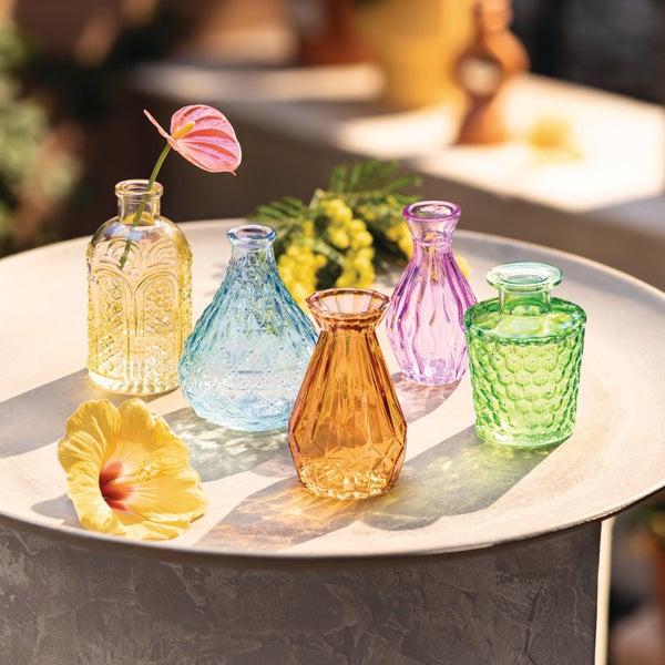 Fünf kleine dekorative Glasvasen in verschiedenen Farben und Formen auf einem hellen Tisch mit Blumen.