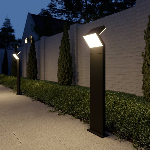 Moderne schwarze LED-Wegeleuchten beleuchten einen gepflasterten Gehweg neben einer Hecke und einer Mauer bei Nacht.