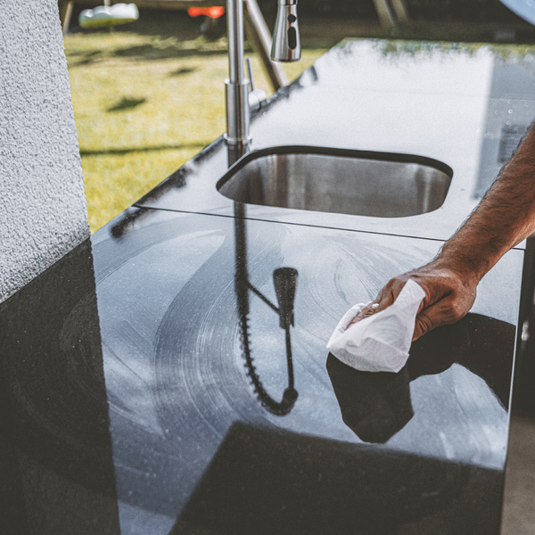 Eine Hand reinigt eine glänzende schwarze Küchenarbeitsplatte mit einem weißen Tuch neben einer Edelstahlspüle.