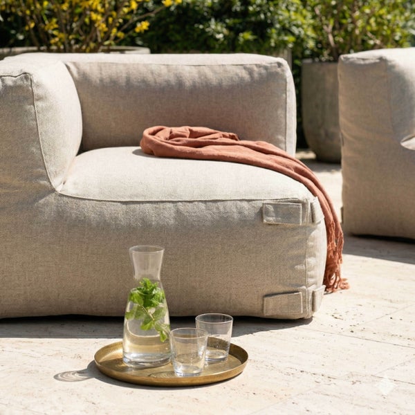Modernes Outdoor-Loungemodul mit hellem Stoffbezug, Plaid und Tablett mit Wasserkaraffe und Gläsern auf einer hellen Terrasse.