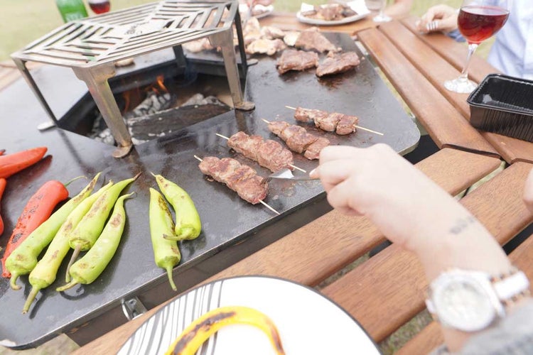 Grilltisch mit integrierter Grillfläche beim Zubereiten von Fleischspießen und Gemüse, umgeben von einem Holztisch in einer geselligen Runde.