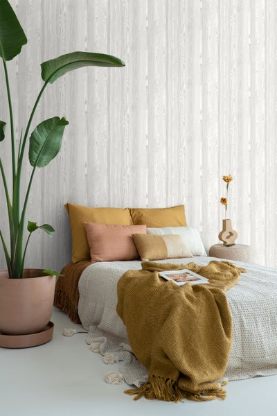 Schlafzimmer mit Bett, Kissen, Decken und einer Zimmerpflanze vor einer Wand mit Holzoptik-Tapete.