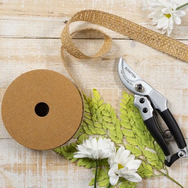 Gartenschere der Marke Schatten aus Karbonstahl mit schwarzen Griffen und eine Rolle Juteband auf einer Holzoberfläche mit Blumen.