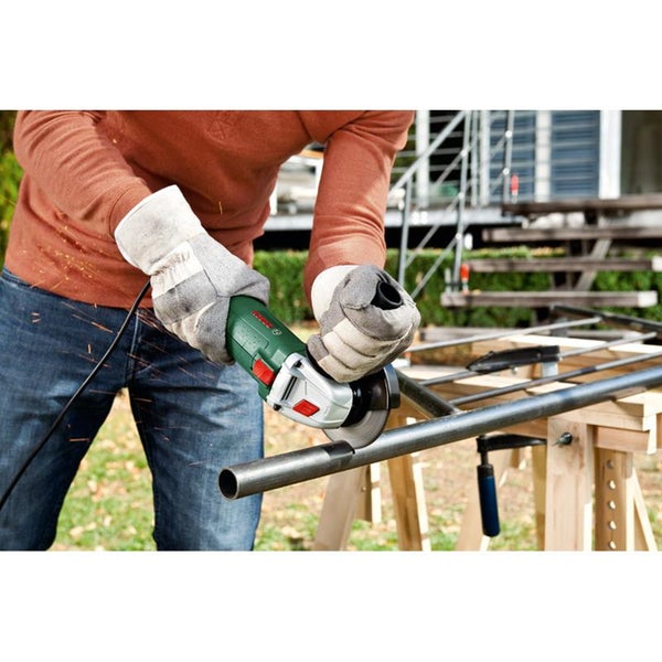 Eine Person benutzt einen Bosch Winkelschleifer, um ein Metallrohr auf einem Holzbock im Garten zu trennen, während sie Schutzhandschuhe trägt.