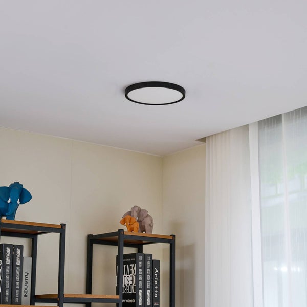 Moderne LED-Deckenleuchte in Schwarz an weißer Decke in einem hellen Wohnbereich mit Regalen und Dekoration.