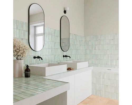 Modernes, helles Badezimmer mit grünen Wandfliesen, zwei weißen Aufsatzwaschbecken, ovalen Spiegeln und schwarzen Wandarmaturen.