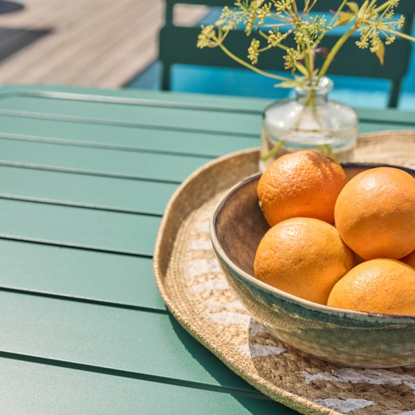 Grüner Gartentisch mit einer Keramikschale voller Orangen auf einem geflochtenen Tablett und einer kleinen Glasvase mit Blumen.