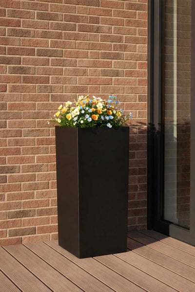 Hoher quadratischer schwarzer Pflanzkübel mit bunten Blumen auf einer Holzterrasse vor einer Ziegelwand.