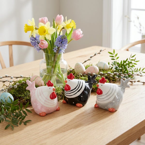 Drei dekorative Keramik Hühnerfiguren als Osterdekoration auf einem Holztisch mit Frühlingsblumen und Ostereiern.