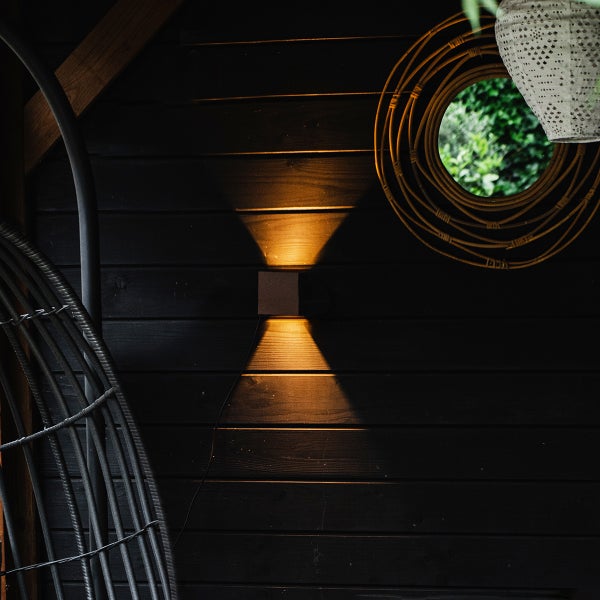 Quadratische Außenwandleuchte mit Lichtkegel nach oben und unten an einer dunklen Holzwand.